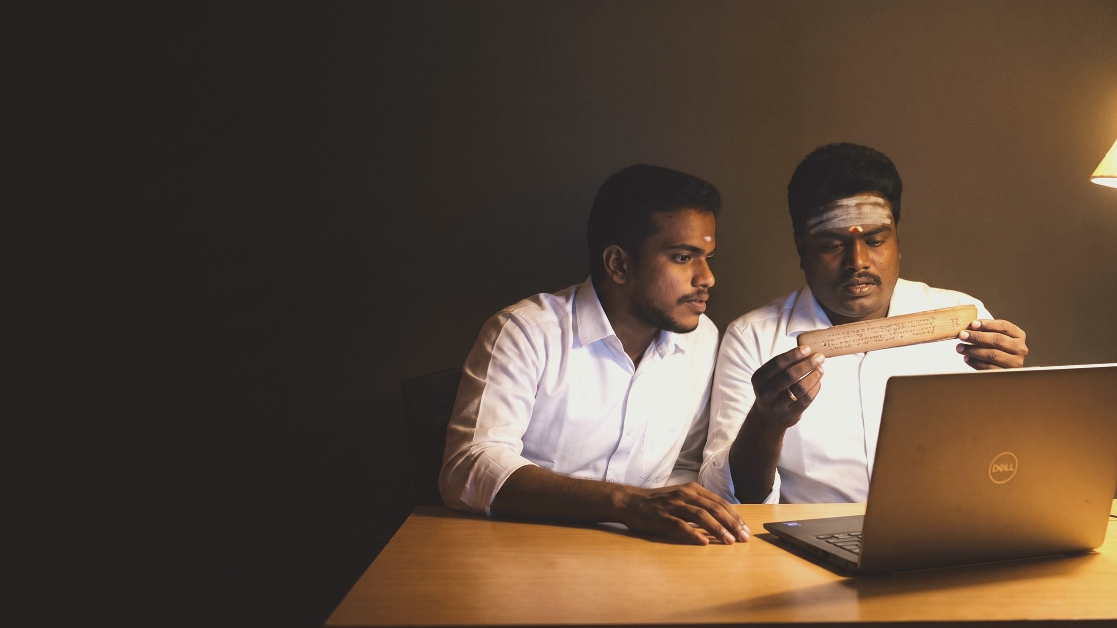 Two readers at the desk, reading a leaf aloud over a laptop