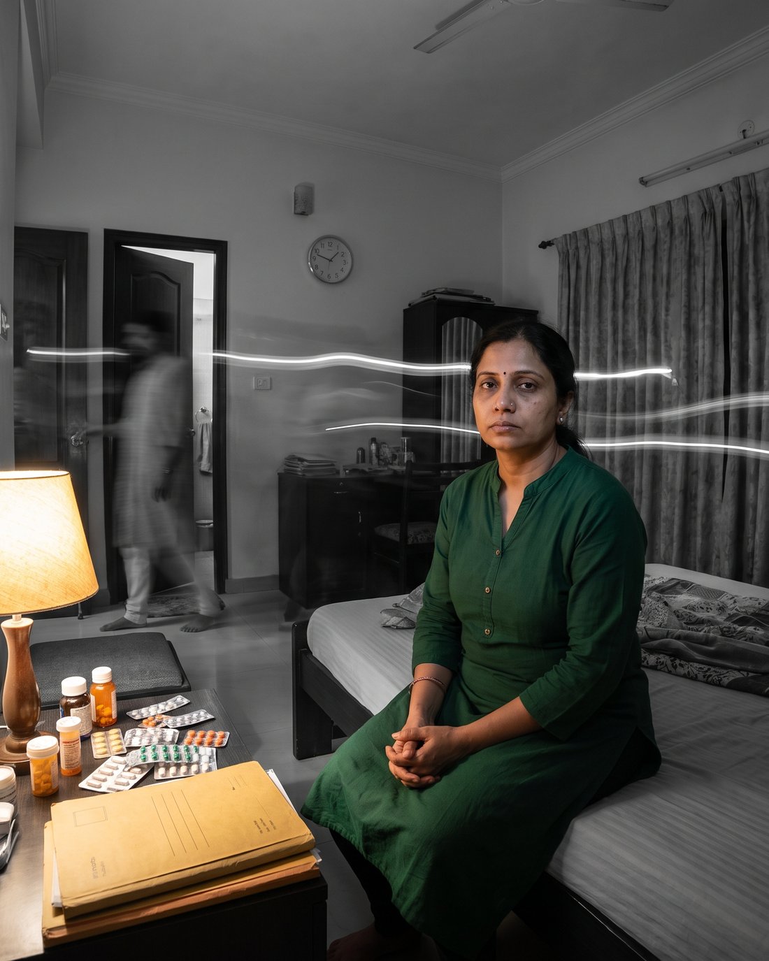 A woman sitting on the edge of a bed, medicines and reports beside her